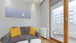 Een moderne woonkamer met een grijze bank, twee mosterdgele kussens, een witte salontafel met een plant, en een schilderij van een vuurtoren aan de muur.