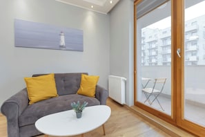 A modern living room with a grey sofa, two mustard-colored cushions, a white coffee table with a plant, and a painting of a lighthouse on the wall.