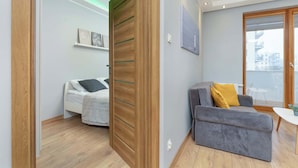 Een moderne woonkamer met een grijze bank, een wit bed en een houten deur met glaspanelen.