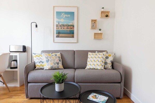 A modern living room with a grey sofa, two patterned cushions, a small round table with a plant, and a framed cityscape poster on the wall. - Dify City - Charpennes (Villeurbanne)