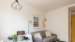 A modern living room with a grey sofa, a dining table set with glassware, a framed picture on the wall, and a geometric light fixture. - Dify City - Charpennes (Villeurbanne)