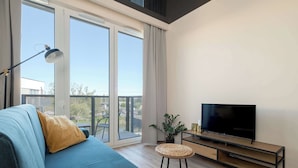 Una sala de estar moderna con un sofá azul, un televisor de pantalla plana sobre un soporte de madera, una lámpara de pie y un balcón con vistas a la ciudad.