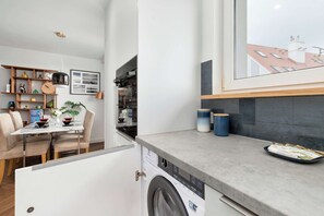 Una cocina compacta con lavadora integrada, un comedor con mesa y sillas, y vistas de la ciudad a través de un ventanal grande.