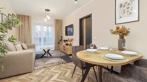 Una sala moderna con un sofá, una mesa de comedor con platos y vasos, un televisor montado en la pared y un ventanal con cortinas.