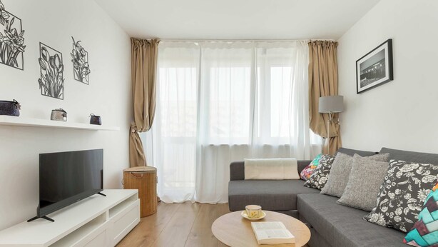 Una sala de estar moderna con un televisor de pantalla plana sobre un mueble blanco, un sofá gris con cojines coloridos, una mesa de centro redonda con un libro y una tetera, y un ventanal con cortinas de gasa.