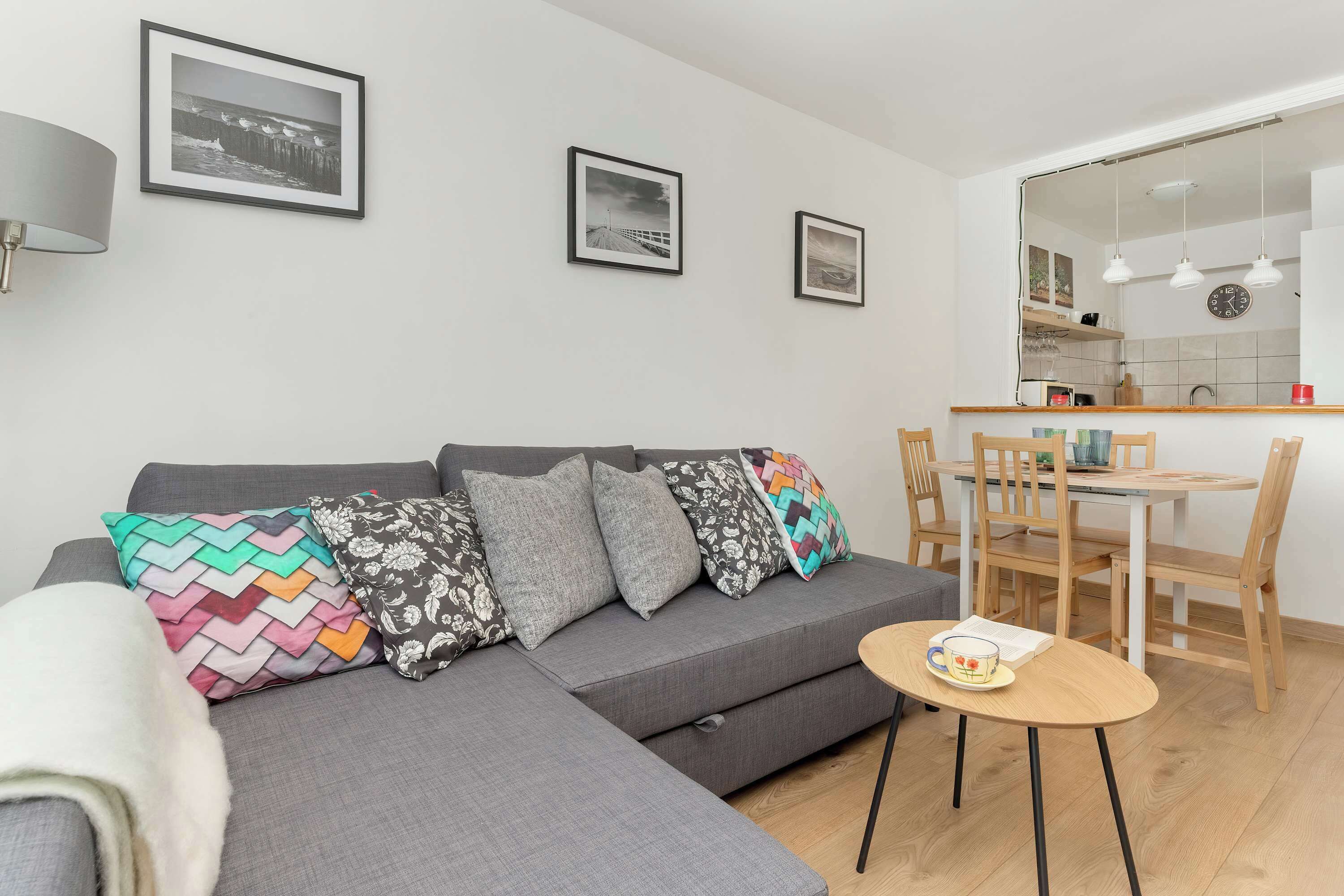 A modern living room with a grey sofa, colorful pillows, a small round table with a mug, and a dining area with wooden chairs and a white table.
