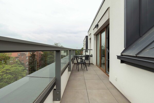 Un balcón con barandilla de vidrio, una mesa con sillas y vistas a edificios y árboles.