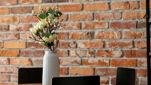 Una mesa con un jarrón blanco de flores, sillas negras y un fondo de pared de ladrillo.