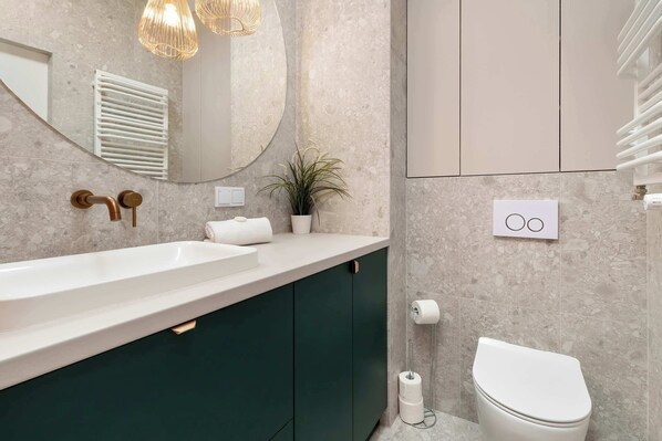 A modern bathroom with a white sink, dark green cabinets, a white toilet, a mirror, a potted plant, and a towel rack.