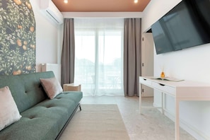 A modern living room with a sofa, a desk, a flat-screen TV, and a view through a large window.