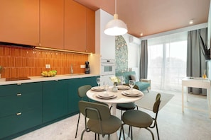 A modern kitchen with teal cabinets, a dining area with a round table, and a living space with a sofa and large windows.