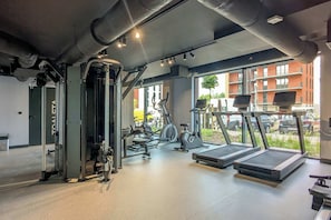 A gym with exercise equipment, a treadmill, and a window with a view of a building and parked cars.