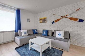 The comfortable gray sofa, decorated with cushions featuring nautical motifs such as steering wheels and sailboats, is an ideal place to relax. On the white brick wall, there is a decoration in the form of wooden oars, which adds charm and character.