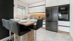 Une cuisine moderne avec des meubles blancs, un réfrigérateur en acier inoxydable et une table à manger en bois, assortie de chaises noires.