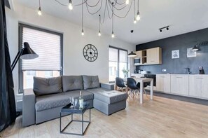 Uma sala de estar moderna com um sofá modular cinza, uma mesa de centro e uma área de refeições com mesa e cadeiras. Há também uma cozinha com armários e micro-ondas.