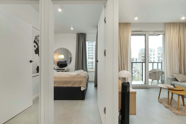 Une chambre d’hôtel moderne comprenant un lit, un coin salon avec une table et des chaises, une grande fenêtre avec des rideaux et un balcon offrant une vue sur la ville.