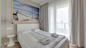 Una habitación de hotel con cama, un cuadro de un barco en la playa, una mesita de noche y una ventana con cortinas.