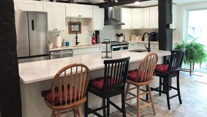 A modern kitchen with white cabinets, a large island with bar stools, a black ceiling beam, and a view of the outdoors.