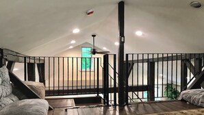 A loft bedroom with a high ceiling, exposed beams, a wooden staircase, a balcony with a metal railing, and a green door leading outside.