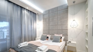 Una habitación de hotel moderna con una cama grande, dos mesitas de noche, una ventana con cortinas y una lámpara colgante de diseño geométrico.