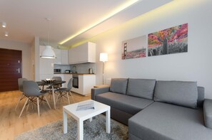 Una sala de estar moderna con un sofá gris, una mesa de comedor con sillas, una mesa de centro blanca, una zona de cocina y un cuadro en la pared.