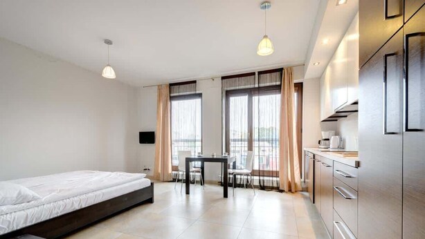 A modern hotel room with a bed, a dining table, and a kitchenette.