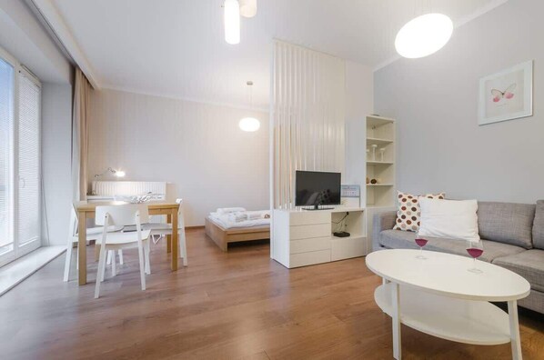 Ein modernes Wohnzimmer mit einer Couch, einem Esstisch mit Stühlen, einem Fernseher und einem kleinen weißen Tisch mit Gläsern.