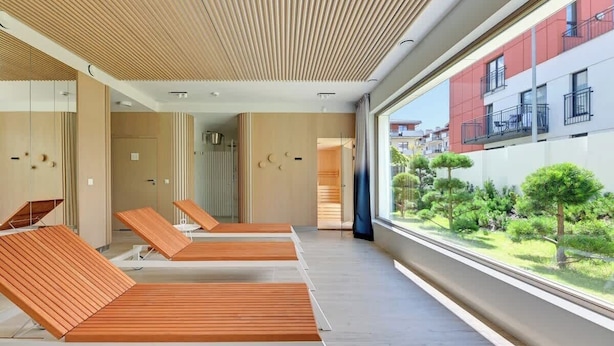 A modern spa area with wooden loungers, a large window with a view of a courtyard, and a ceiling with a wooden slat design.