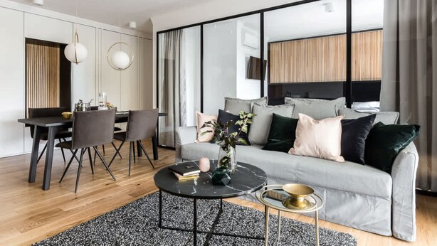 A modern living room with a grey sofa, a dining table with chairs, a coffee table, and a large window with curtains.