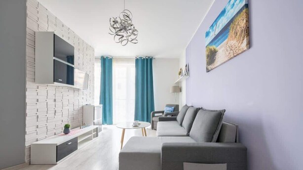 A modern living room with a grey sofa, a flat-screen TV mounted on a white brick wall, a small table with a plant, and a painting of a beach scene on the wall.