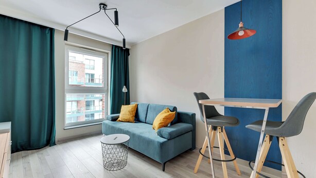 A modern living room with a blue sofa, two yellow cushions, a round coffee table, and a small side table. There is a window with curtains, a blue accent wall, and a red pendant light. - Comfortable Apartment in the Center | Remote Work (Gdańsk)