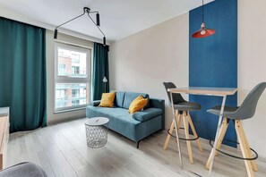 A modern living room with a blue sofa, two yellow cushions, a round coffee table, and a small side table. There is a window with curtains, a blue accent wall, and a red pendant light. - Comfortable Apartment in the Center | Remote Work (Gdańsk)
