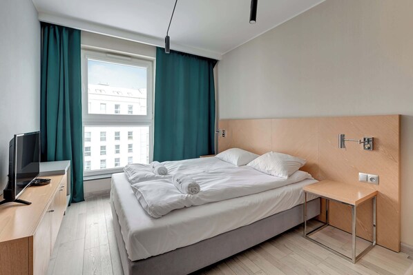A modern hotel room with a bed, a TV, a desk, and a window with curtains.