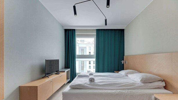 Ein modernes Schlafzimmer mit einem Bett, einem Fernseher auf einer Holzeinrichtung und Blick auf Gebäude durch ein Fenster.