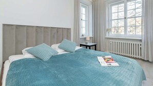 Un lit bien fait, recouvert d’une couvre-lit turquoise et agrémenté d’oreillers, une table de chevet avec une lampe, et une fenêtre avec des rideaux.