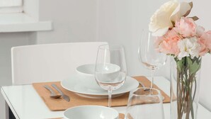 Une table à manger dressée avec de l’assiette blanche, des bols, des couverts et des verres à vin, agrémentée d’un vase de fleurs.