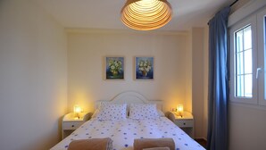 A neatly made bed with floral bedding, two wall-mounted paintings, bedside lamps, and a window with curtains. - La Marina 1 5 - Two Bedroom Apartment, Sleeps 5 (Punta del moral)