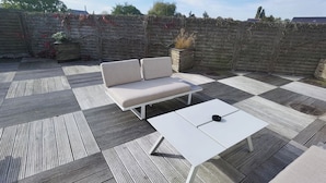 A patio with a beige sofa and a white table, surrounded by a wooden deck and a stone wall. - Relax Gent Wellness (Lievegem)