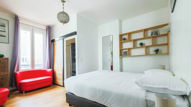 Une chambre moderne avec un grand lit, un canapé rouge, une étagère en bois remplie de livres et une grande fenêtre avec des rideaux.