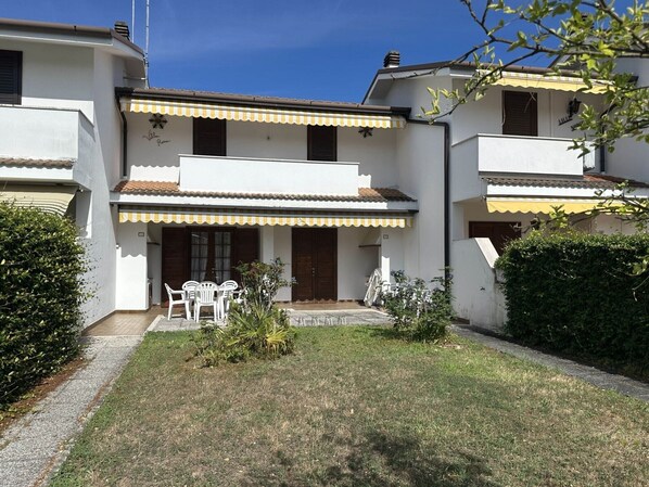 Un edificio residencial de dos plantas con un pequeño jardín delantero. Cuenta con dos balcones con toldos, una puerta y un patio pequeño con mesa y sillas.