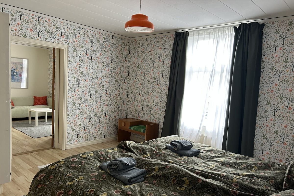 A bedroom with floral wallpaper, a bed with a floral bedspread, a small wooden nightstand, a white table, and a view of another room through an open door.