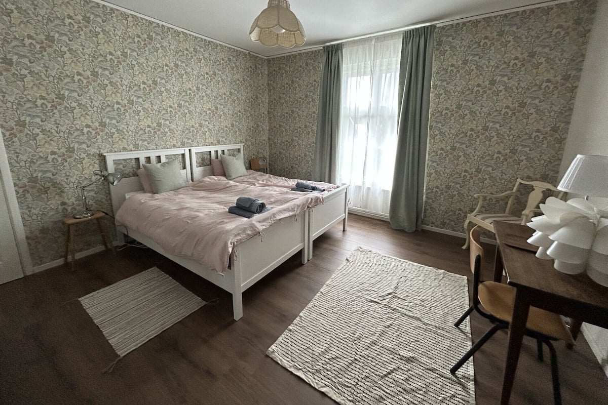 A bedroom with a bed, a desk, a chair, a lamp, and a hanging light fixture.