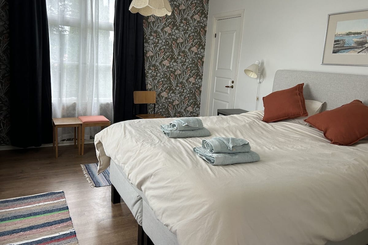A neatly made bed with rolled towels, a wooden chair, a desk, and a lamp.