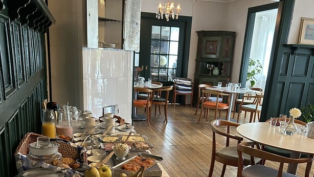 Un buffet de petit-déjeuner proposant divers plats et couverts, des chaises en bois et un lustre, dans une pièce au sol en bois et aux murs verts.