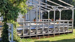 Un espace extérieur aménagé, abrité et doté de bancs et de tables en bois, entouré de plantes en pot et d’une clôture en bois.