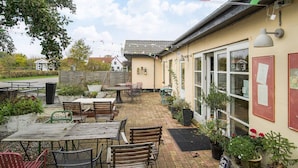 An outdoor patio area with tables and chairs, potted plants, and a building with large windows and a satellite dish. - 2 Person Holiday Park Home in Gorlev (Gørlev)