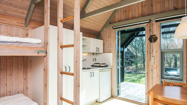 A wooden cabin interior with a bunk bed, kitchenette, and a dining area with a view of the outdoors. - 6 Person Holiday Park Home in Pandrup (Pandrup)