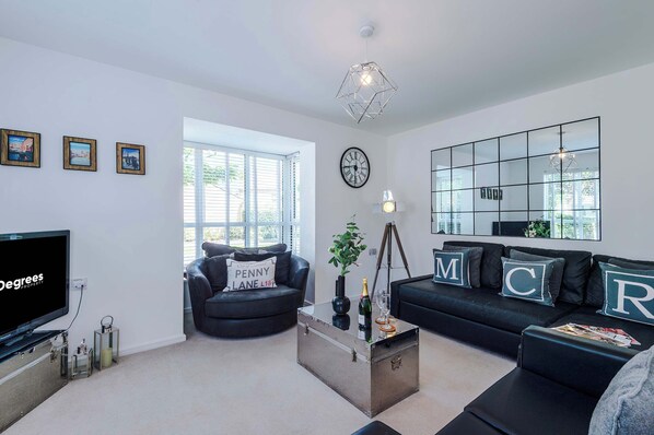 A modern living room with a large flat-screen TV, a black leather sofa, a coffee table, and a large window providing natural light.
