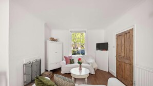 Une petite pièce aux murs blancs, avec une porte en bois, un canapé blanc agrémenté de coussins, une petite table blanche et un radiateur.