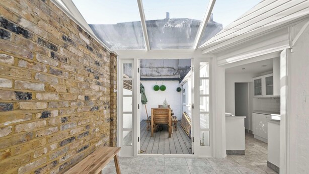 Un couloir étroit mène à un banc en bois et à une table. Un mur de briques se trouve d’un côté et une cuisine blanche de l’autre.
